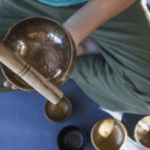 一位穿著橄欖綠褲子的人盤腿坐在瑜珈墊上，手中拿著一個頌缽和缽槌，地上還有其他幾個頌缽