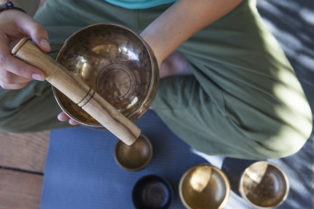 一位穿著橄欖綠褲子的人盤腿坐在瑜珈墊上，手中拿著一個頌缽和缽槌，地上還有其他幾個頌缽