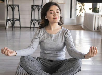一位女性穿著灰色居家服，戴著無線耳機，盤腿坐在地板上冥想，雙手做出禪定手印