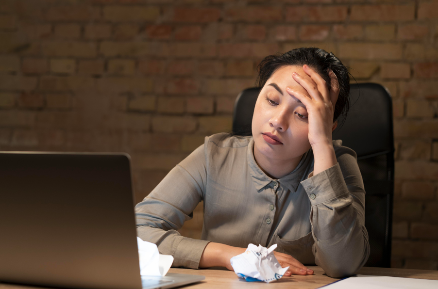 一位表情痛苦的女性坐在辦公桌前，一隻手撐著額頭，桌上有筆記型電腦和揉成一團的紙張，背景是磚牆