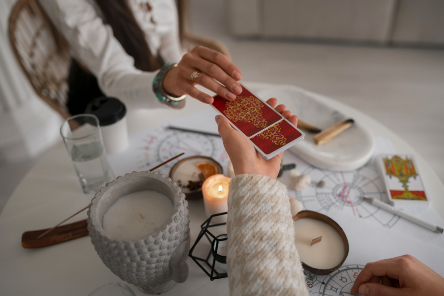 兩隻手正在傳遞一副背面印有紅色圖案的卡牌，背景桌面散落著蠟燭、香、水杯以及占星圖