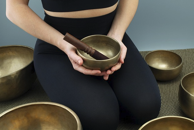 一位穿著黑色運動服的女性跪坐在墊子上，雙手捧著一個頌缽，缽內放著一個深色缽槌，周圍還散落著其他頌缽