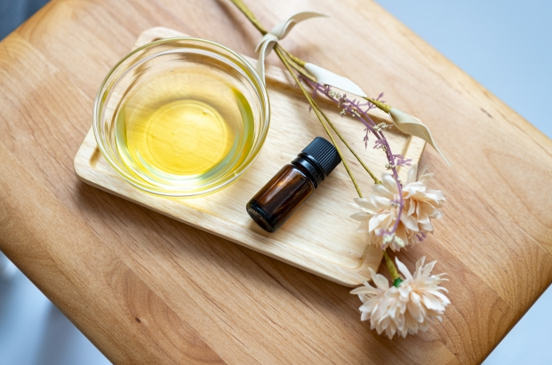 一個裝有透明黃色液體的玻璃碗和一瓶深色精油瓶，與幾朵乾燥的花束一同放在木製托盤上