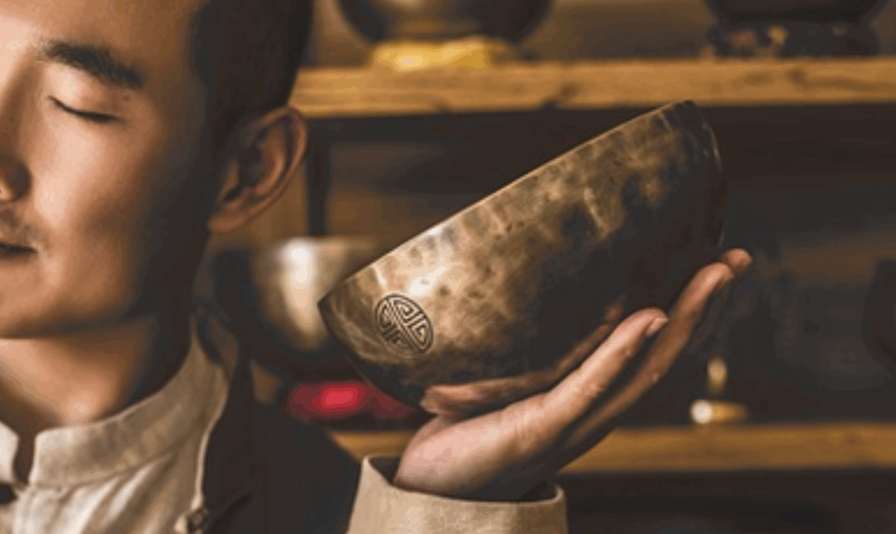 一位閉著眼睛的男性，將一個刻有圖案的銅製頌缽靠近臉部，背景是擺滿頌缽的木製層架