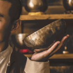 一位閉著眼睛的男性，將一個刻有圖案的銅製頌缽靠近臉部，背景是擺滿頌缽的木製層架