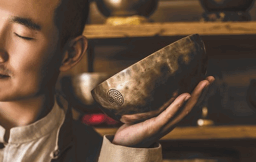 一位閉著眼睛的男性，將一個刻有圖案的銅製頌缽靠近臉部，背景是擺滿頌缽的木製層架