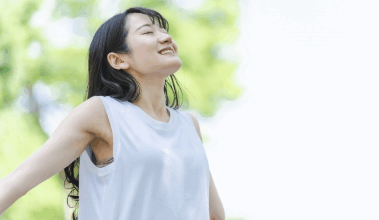 一位亞洲女性穿著白色無袖上衣，閉著眼睛，抬頭享受陽光和戶外清新的空氣，背景是模糊的綠色樹葉