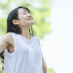 一位亞洲女性穿著白色無袖上衣，閉著眼睛，抬頭享受陽光和戶外清新的空氣，背景是模糊的綠色樹葉