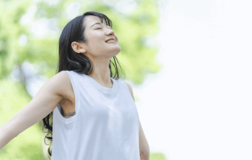 一位亞洲女性穿著白色無袖上衣，閉著眼睛，抬頭享受陽光和戶外清新的空氣，背景是模糊的綠色樹葉