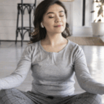 一位女性穿著灰色居家服，戴著無線耳機，盤腿坐在地板上冥想，雙手做出禪定手印。