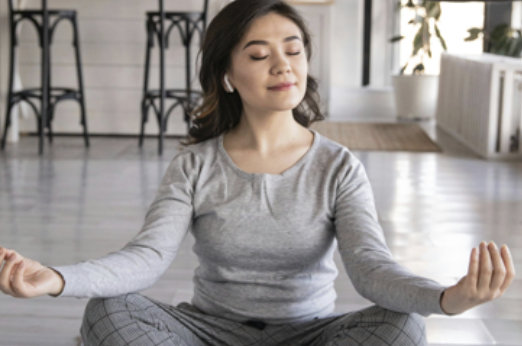 一位女性穿著灰色居家服，戴著無線耳機，盤腿坐在地板上冥想，雙手做出禪定手印。