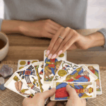 兩雙手在木桌上操作塔羅牌，桌面上擺有馬賽塔羅牌、茶杯、水晶和一塊粗麻布墊