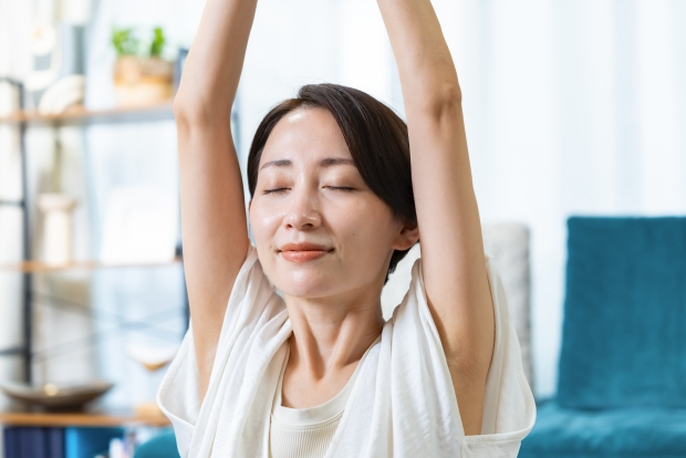 一位中年女性閉著眼睛，在明亮的客廳中進行伸展靜坐，雙手高舉過頭，表情平靜放鬆。