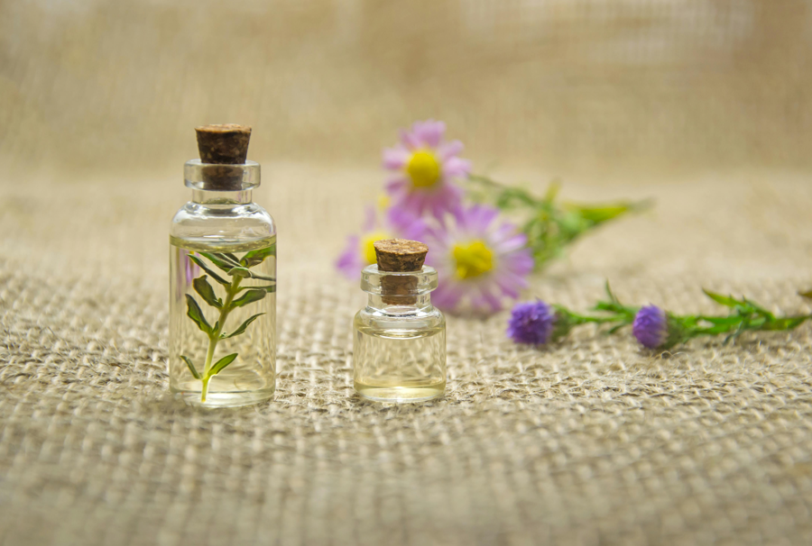 兩個軟木塞精油瓶和草本植物，放置在粗麻布背景上，背景虛化處有粉紫色小花裝飾