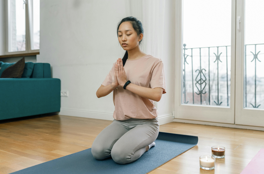 一位年輕女性在室內瑜珈墊上，閉眼跪坐並雙手合十進行冥想，背景有窗戶、陽台門和點燃的香氛蠟燭，展現居家身心放鬆的氛圍