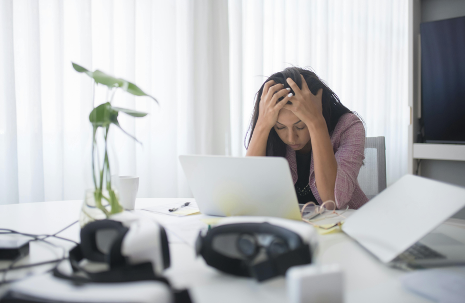 一位身穿粉色西裝外套的女性坐在辦公桌前，用雙手抱著頭，看起來非常沮喪、壓力大或精疲力盡，桌上放著筆記型電腦、眼鏡和VR頭戴裝置，背景是明亮的白色窗簾