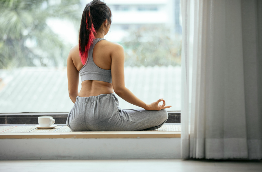 一位年輕女性背對鏡頭，紮著有粉紅色挑染的馬尾，穿著灰色運動內衣和長褲，在窗邊的墊子上盤腿坐著進行冥想，右手做出智慧手印，旁邊放著一杯熱飲