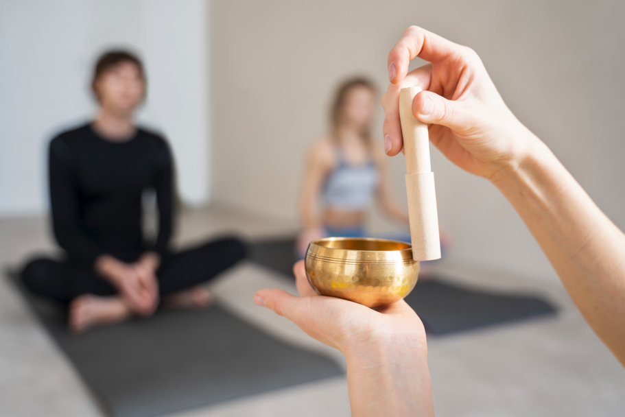 特寫鏡頭顯示一隻手拿著小型銅製頌缽和木槌，準備在冥想休息，背景有兩位模糊的人影盤腿坐在瑜珈墊上休息或冥想