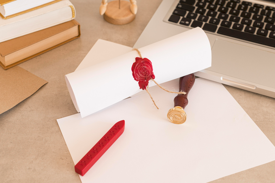 一張捲起來的文件或證書，用麻繩和紅色蠟封蓋章，旁邊擺放著木柄印章、紅色火漆蠟條、筆記型電腦和一疊書，呈現學術成就的主題
