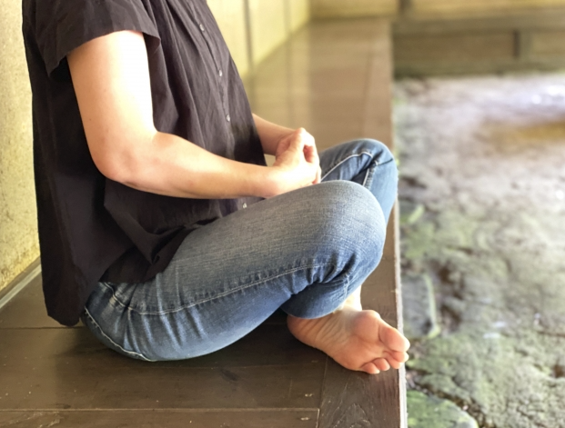 一個人赤腳穿著牛仔褲和深色上衣，盤腿坐在木質台階上，在庭院中靜坐，突顯放鬆和專注。