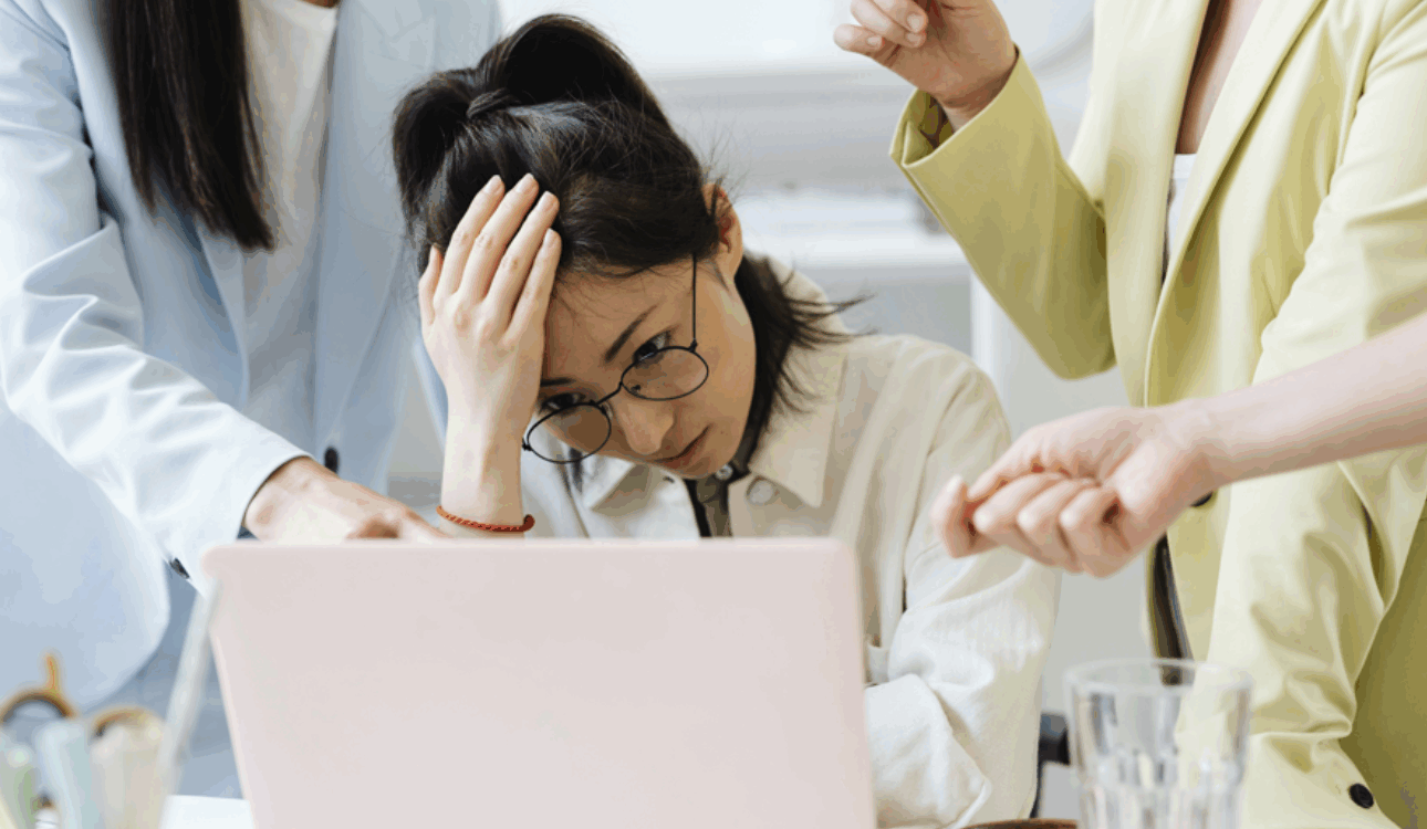 一名戴眼鏡的女性員工在筆記型電腦前抱著頭，看起來壓力大及焦慮，兩位穿著西裝外套的同事或主管在旁指責或施加壓力，描繪職場壓力、倦怠或衝突的情境