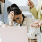 一名戴眼鏡的女性員工在筆記型電腦前抱著頭，看起來壓力大及焦慮，兩位穿著西裝外套的同事或主管在旁指責或施加壓力，描繪職場壓力、倦怠或衝突的情境