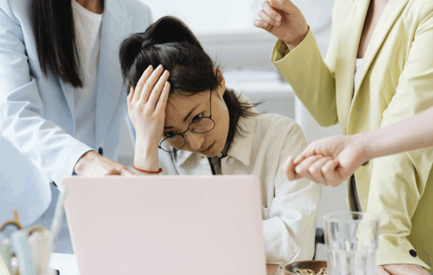 一名戴眼鏡的女性員工在筆記型電腦前抱著頭，看起來壓力大及焦慮，兩位穿著西裝外套的同事或主管在旁指責或施加壓力，描繪職場壓力、倦怠或衝突的情境