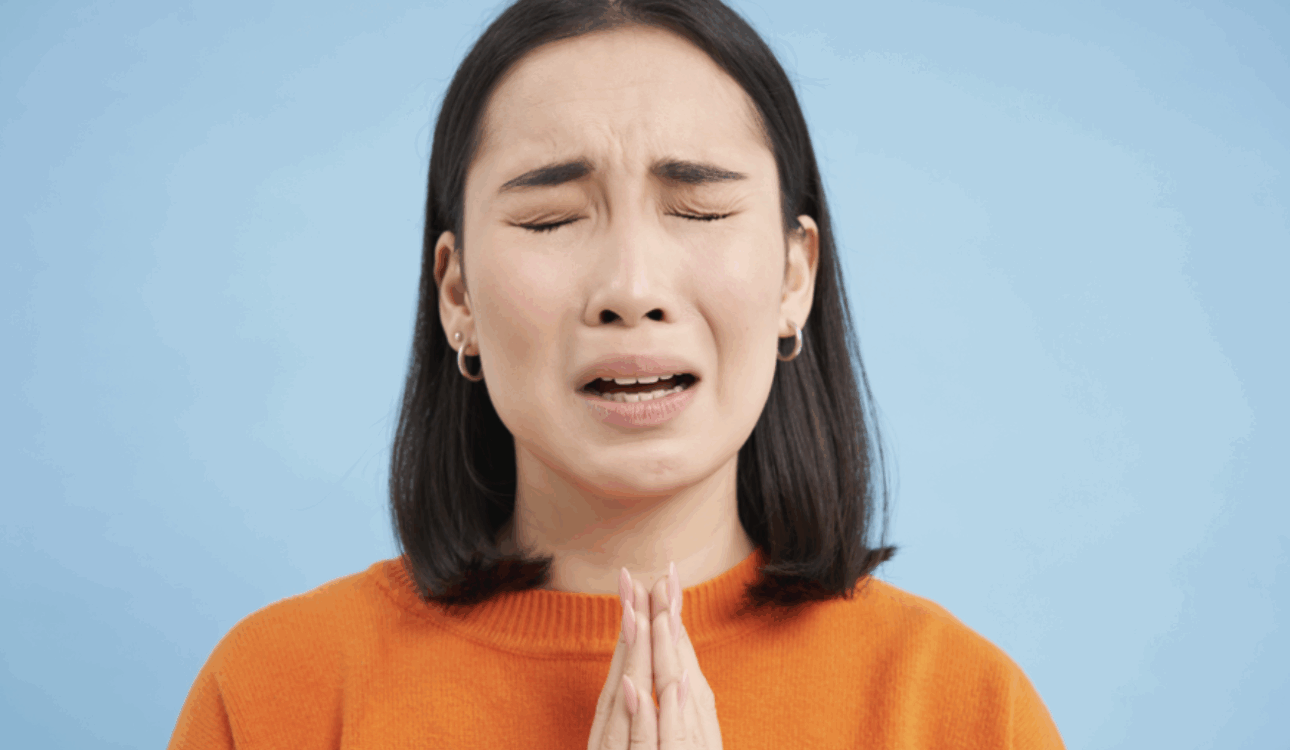 悲傷的年輕女性，緊閉雙眼，雙手合十祈禱或懇求幫助，身穿橘色上衣，背景為藍色