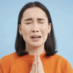 悲傷的年輕女性，緊閉雙眼，雙手合十祈禱或懇求幫助，身穿橘色上衣，背景為藍色