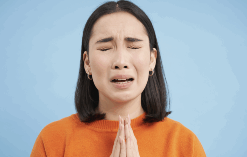 悲傷的年輕女性，緊閉雙眼，雙手合十祈禱或懇求幫助，身穿橘色上衣，背景為藍色