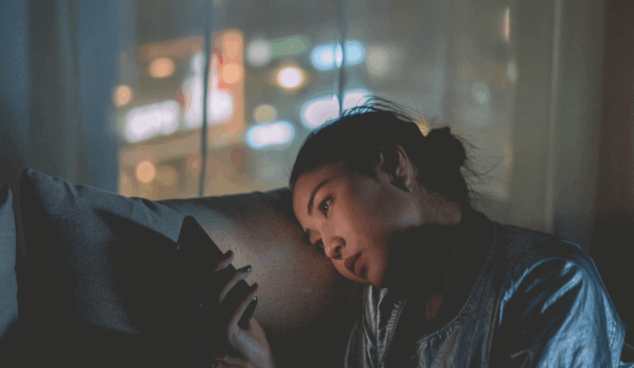 一位年輕女性在昏暗的房間裡，疲憊地斜靠在沙發上，低頭看著手中的智慧型手機，背景是模糊的城市夜景燈光