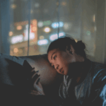 一位年輕女性在昏暗的房間裡，疲憊地斜靠在沙發上，低頭看著手中的智慧型手機，背景是模糊的城市夜景燈光