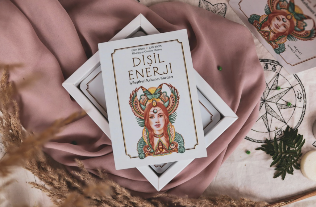 一本名為「DİŞİL ENERJİ」（女性能量）的書籍放在白色方框中，封面印有一名帶著華麗頭飾的女性插畫。背景裝飾著粉紅色絲綢、乾燥花、占星羅盤圖案的布料以及散落的綠色小珠子
