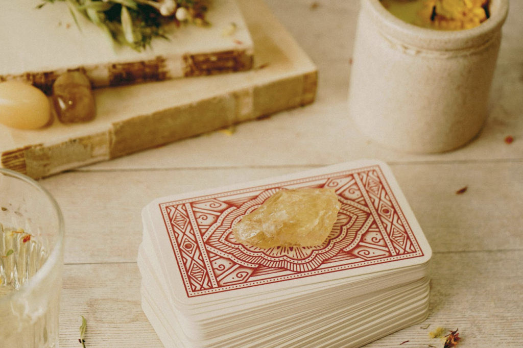  木質桌面上疊放著一疊背面為紅底白花紋的塔羅牌，牌組上方放置著一顆半透明的黃水晶原石。背景可見乾燥花束、書籍、水晶以及一個裝有黃色花朵的香氛容器。