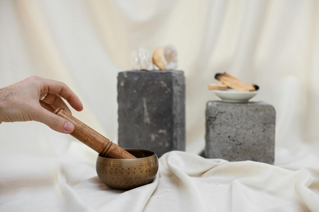 一隻手正握著木槌敲擊一個銅製的頌缽（Singing Bowl），擺放在柔和的米白色布景上，背景有兩塊石塊，上面分別擺放著水晶原石和聖木（Palo Santo），展現冥想、聲音療癒、靈性儀式或淨化空間的主題。