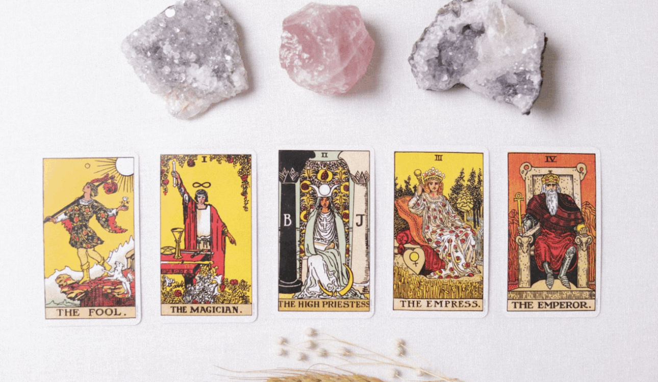 一組塔羅牌主要牌組（愚者、魔術師、女祭司、皇后、皇帝）整齊地排列在白色表面上，上方擺放著三塊天然水晶或礦石（兩塊白色的晶洞與一塊粉色的薔薇石），下方點綴著麥穗和乾燥的穀物，營造出神祕學、占卜、身心靈或能量療癒的主題。