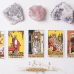 一組塔羅牌主要牌組（愚者、魔術師、女祭司、皇后、皇帝）整齊地排列在白色表面上，上方擺放著三塊天然水晶或礦石（兩塊白色的晶洞與一塊粉色的薔薇石），下方點綴著麥穗和乾燥的穀物，營造出神祕學、占卜、身心靈或能量療癒的主題。