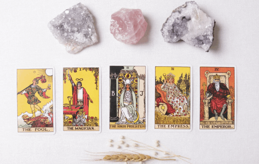 一組塔羅牌主要牌組（愚者、魔術師、女祭司、皇后、皇帝）整齊地排列在白色表面上，上方擺放著三塊天然水晶或礦石（兩塊白色的晶洞與一塊粉色的薔薇石），下方點綴著麥穗和乾燥的穀物，營造出神祕學、占卜、身心靈或能量療癒的主題。