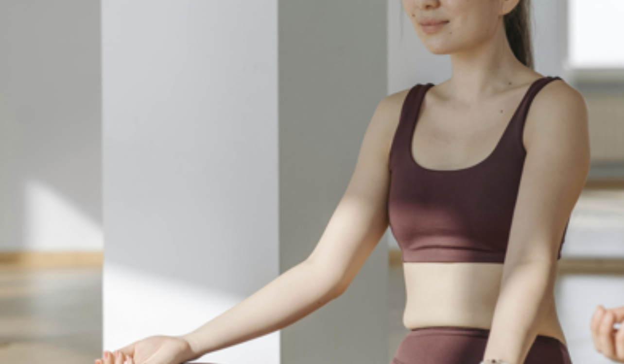 一名亞裔女性穿著深紅褐色運動背心與緊身褲，閉著雙眼以蓮花坐姿在木質地板上進行冥想。她的前方放著兩塊密佈細針的釘床木板（Sadhu board），背景為明亮且簡約的室內瑜珈工作室