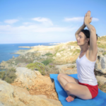 一名穿著白色背心與紅色短褲的女性，坐在藍色瑜珈墊上，於壯麗的海邊岩石頂端進行瑜珈。她閉著雙眼，雙手合十高舉過頭（樹式或坐姿伸展），背景是湛藍的大海、蜿蜒的海岸線與晴朗的天空。