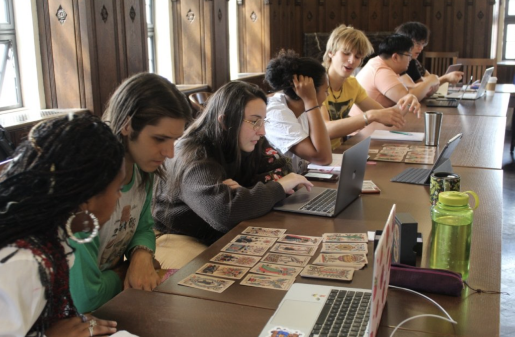 一群年輕人圍坐長桌前進行塔羅牌學習與討論，結合筆電與牌卡，呈現互動式的學習場景。