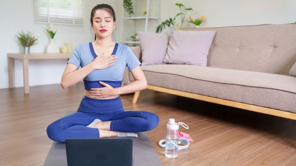女性在居家木地板上跟著筆電進行瑜珈練習，雙手放在胸口與腹部感受呼吸。