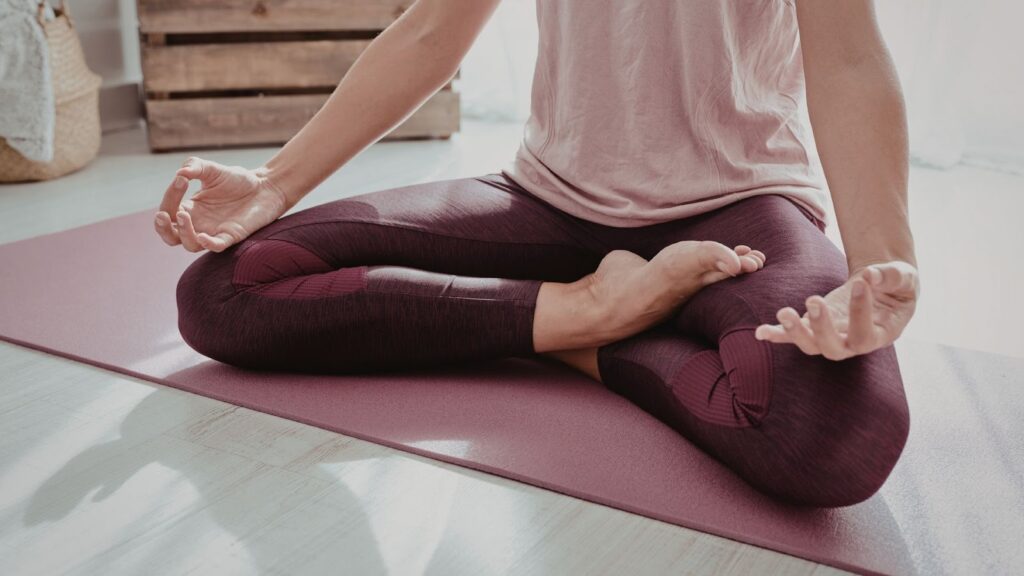 瑜伽墊上的盤腿坐姿特寫，展示標準的蓮花坐姿與放鬆的手部動作，光影柔和。