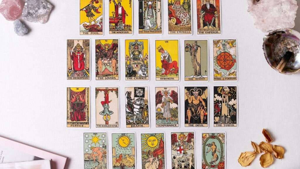 在純白背景上，塔羅牌的大阿爾克那（Major Arcana）牌組呈三排整齊排列，周圍點綴著粉晶、紫水晶原石、貝殼香爐以及散落的乾燥花瓣，呈現出明亮且純淨的神祕學視覺風格。
