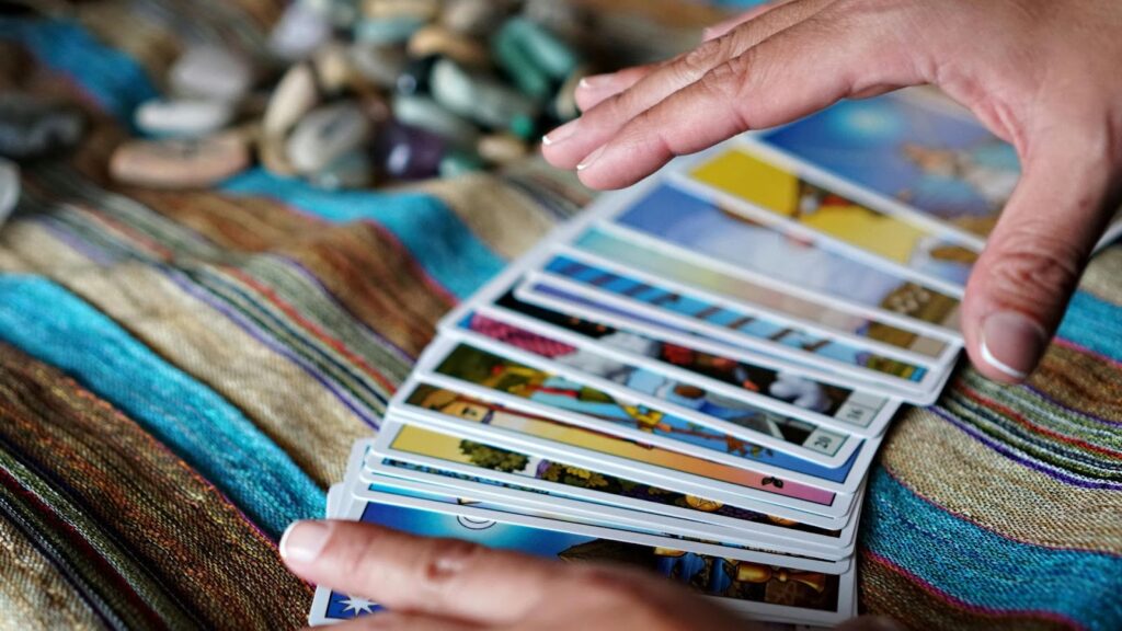 在色彩繽紛的條紋布面上，一名占卜師的雙手正拂過一疊呈扇形展開的塔羅牌，背景有一堆天然原石，展現出直覺感應與神祕占卜的過程