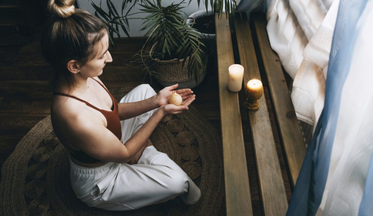 女子在燭光與綠植環繞的空間中盤坐冥想，雙手捧著物件，營造安定而內省的靜心時刻