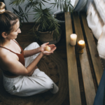 女子在燭光與綠植環繞的空間中盤坐冥想，雙手捧著物件，營造安定而內省的靜心時刻