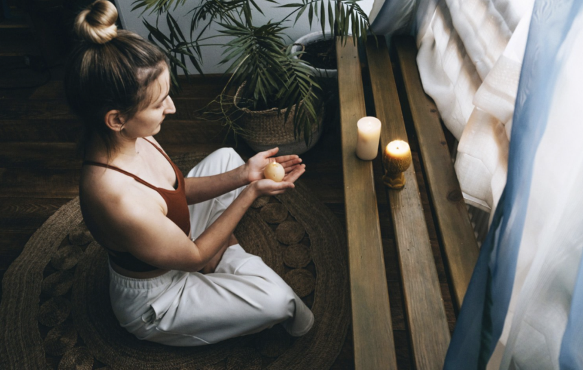 女子在燭光與綠植環繞的空間中盤坐冥想，雙手捧著物件，營造安定而內省的靜心時刻