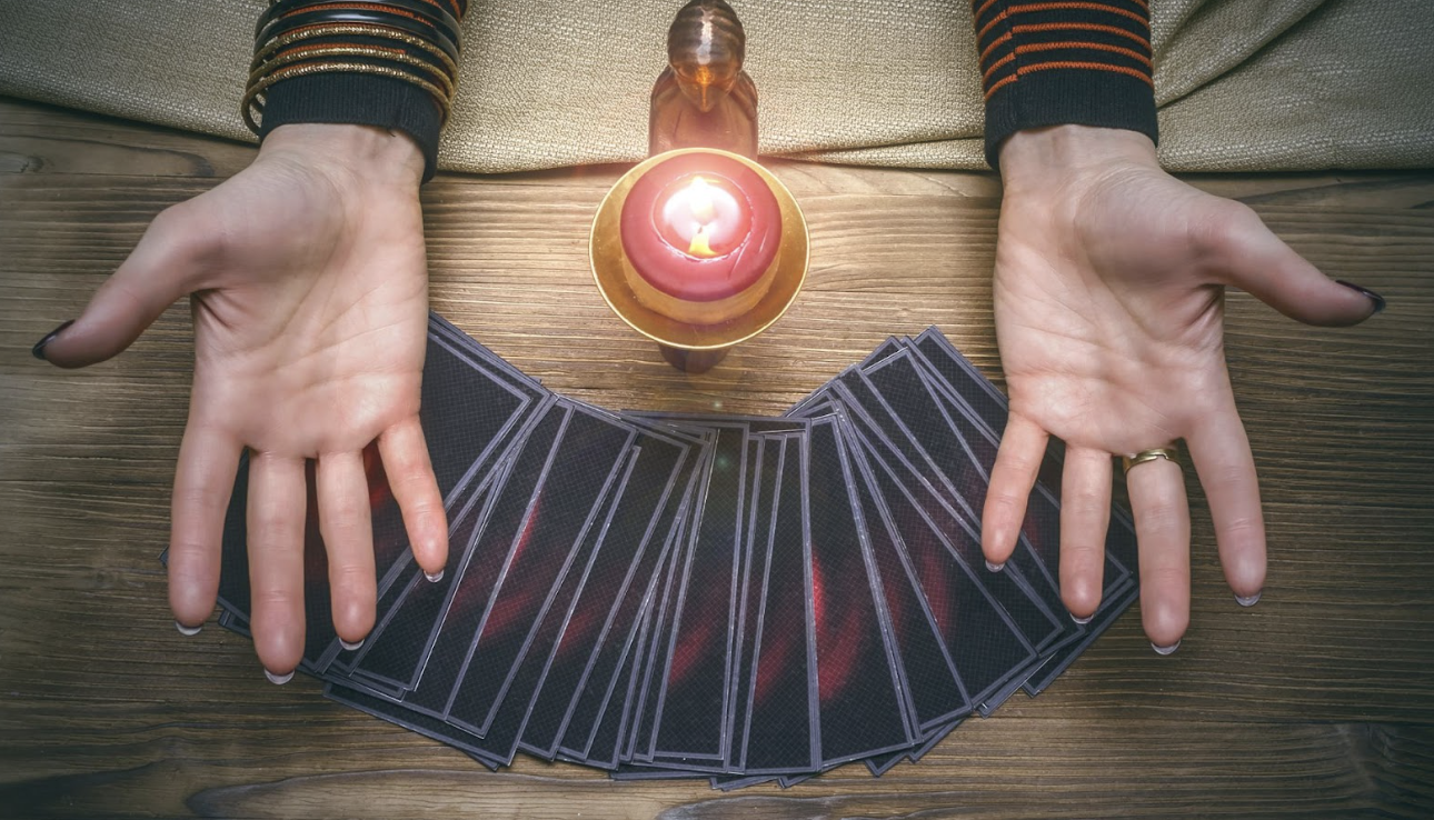 在木質桌面上，一名占卜師張開雙手展示前方呈扇形排列的塔羅牌，中間點燃著一顆紅色蠟燭，營造出神祕且充滿靈性指引的氛圍。