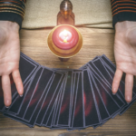 在木質桌面上，一名占卜師張開雙手展示前方呈扇形排列的塔羅牌，中間點燃著一顆紅色蠟燭，營造出神祕且充滿靈性指引的氛圍。